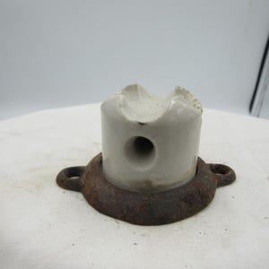 May include: A vintage electrical insulator, featuring a cylindrical ceramic body with a central hole and a chipped top. It is mounted on a circular, rusty metal base with two loop attachments. The insulator is off-white.