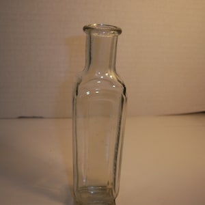 May include: A clear glass bottle with a square shape and a rounded top. The bottle is empty and has a smooth surface.