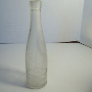 May include: A clear glass bottle with a long, slender neck and a rounded body. The bottle has a slightly textured surface and appears to be empty. The bottle has a label with text that is difficult to read. The bottle is likely vintage.