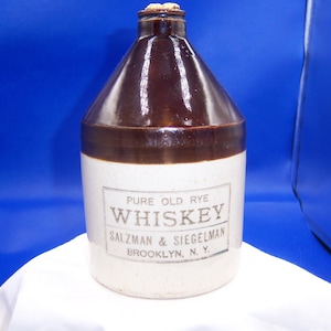 May include: A vintage whiskey jug with a brown glazed top and a cream-colored base. The jug features a label with the text "PURE OLD RYE WHISKEY SALZMAN & SIEGELMAN BROOKLYN, N.Y." The jug is sealed with a cork stopper.