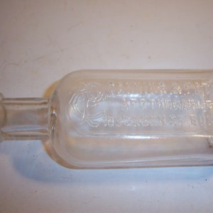 May include: A clear glass bottle with a textured surface. The bottle has a label that reads "D.C. Weimar & Bro. Apothecaries, 103 Green St. Bklyn."