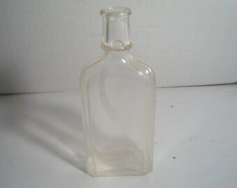 May include: A clear, rectangular glass bottle with a narrow neck and a rounded shoulder. The bottle has vertical ridges and a slightly curved shape. The bottle is empty and appears to be vintage.