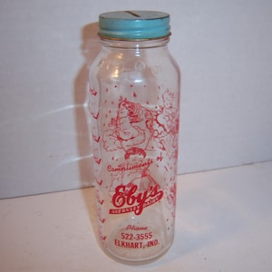 May include: A clear glass milk bottle with a blue lid. The bottle has a red and white design with the text "Compliments of Eby's Guernsey Dairy Phone 522-3555 Elkhart, Ind."