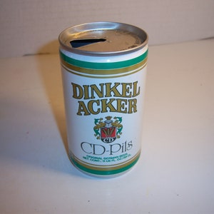 May include: A white aluminum can of Dinkel Acker CD-Pils Original German Beer. The can has a green and gold label with a crest and the text "Dinkel Acker CD-Pils Original German Beer Net Cont. 11 US Fl. Oz. 325 ml."