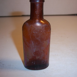 May include: A small, amber-colored glass bottle with a rounded bottom and a narrow neck. The bottle is slightly frosted and has a few scratches.