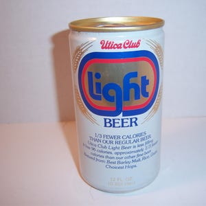 May include: A silver can of Utica Club Light Beer. The can has a gold top and a blue and red label with the words "Utica Club" and "Light" in gold lettering. The can also has the text "1/3 Fewer Calories Than Our Regular Beer" and "12 fl oz (355 ml)".