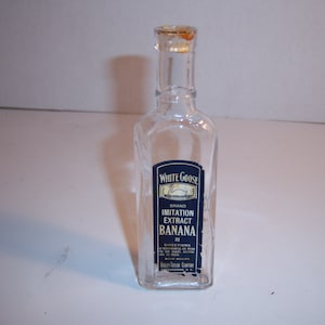 May include: Clear glass bottle with a cork stopper. The bottle has a rectangular label with the text "White Goose Brand Imitation Extract Banana." The label is dark blue with white text and a white goose illustration.