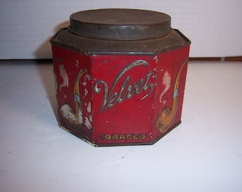 Può includere: Scatola ottagonale rossa con un coperchio di metallo. La scatola ha un'etichetta bianca con il testo "Velvet Tobacco" e un'immagine stilizzata di una pipa.
