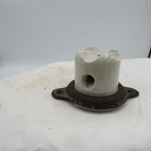 May include: A cylindrical, off-white ceramic object with a circular hole and a damaged top, mounted on a rusty, circular metal base with two mounting holes. The object is set against a white background.