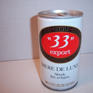 May include: A white aluminum can of "33" export beer. The can has a red and gold label with the text "33" in a large font and the words "export" and "Biere de Luxe" in smaller font. The label also includes the text "blonde fine et legere" and "Union de Brasseries".