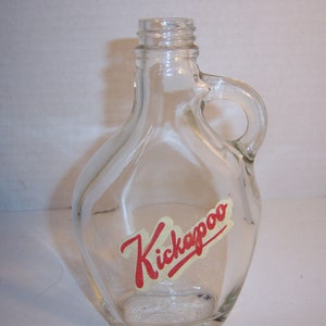 May include: Clear glass bottle with a handle and the word "Kickapoo" in red script on a yellow banner. The bottle has a rounded shape and a small opening at the top.