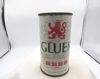 Könnte beinhalten: Vintage Gluck Fine Pilsner Bierdose. Die zylindrische Dose hat einen weißen Hintergrund und ein rotes Löwenemblem. Das Wort "GLUEK" ist in Schwarz gedruckt, darunter "Fine Pilsner Beer".