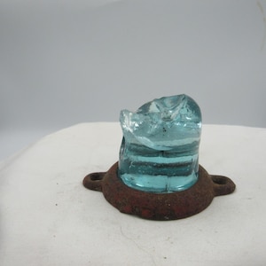 May include: A vintage, light blue glass insulator with a chipped top, mounted on a rusty, circular metal base. The insulator has horizontal ridges and a clear, textured appearance. The base has two small loops.