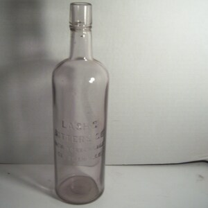 May include: A clear, antique bottle with a light purple tint. The bottle has a long neck and a cylindrical body. The text "LASH'S BITTERS CO. NEW YORK, N.Y." is embossed on the front. The bottle is approximately 10 inches tall.