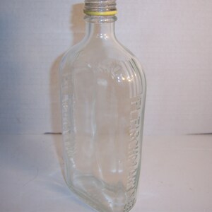May include: Clear glass bottle with a flat, rectangular body and a rounded top. The bottle has a metal cap with a yellow ring. The words "FLEISCHMANN" are embossed vertically on the side of the bottle.