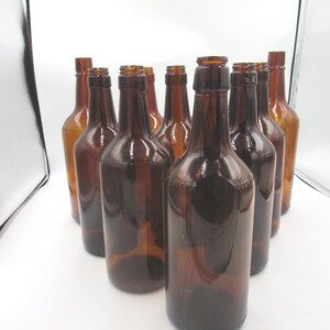 May include: A collection of ten empty amber glass bottles arranged in a triangular formation. Each bottle features a slender neck, a rounded shoulder, and a cylindrical body with a smooth finish. The glass is translucent brown throughout.