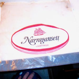 May include: A white plastic bag with a red and white oval logo that says "Narragansett Lager Beer" with a ship illustration inside the oval.