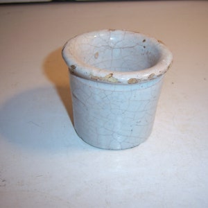 May include: A small, white ceramic pot with a crackled glaze. The pot has a slightly rounded shape and a wide opening.