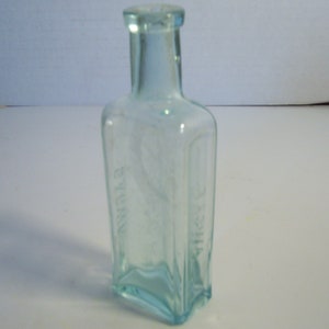 May include: A vintage, rectangular, light blue glass bottle. The bottle has a square base and a rounded top. The words "EYE" and "WASH" are vertically printed on the bottle. The bottle is empty and appears to be an antique.