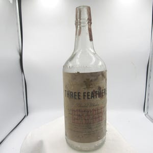 May include: A clear glass bottle with a long neck and a weathered, tan paper label. The label features the text "THREE FEATHERS" in bold black letters, "Blended Whiskey" in script, and a small emblem of three feathers at the top center.