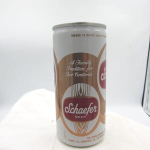 May include: A vintage Schaefer Beer can with a white and gold design. The can features the text "A Family Tradition for Two Centuries" and the Schaefer Beer logo in red. The can is cylindrical and has a silver top.