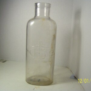 May include: A clear, vintage glass bottle with a rounded rectangular shape. The bottle has a narrow neck and a slightly raised embossed design. The bottle is aged, with some discoloration and wear visible on the surface.