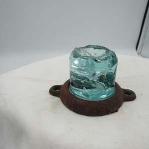 May include: A vintage, light blue glass insulator with a rough, textured top. It sits on a rusty, circular metal base with two loop handles. The glass has air bubbles and imperfections.