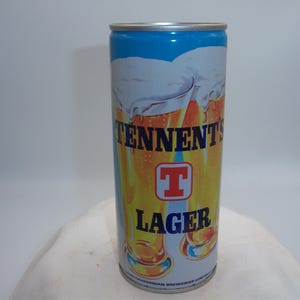 May include: A vintage Tennent's Lager beer can. The can features a blue top, a white foamy beer design, and the words "TENNENT'S LAGER" in blue and orange. A red "T" logo is centered on the can.