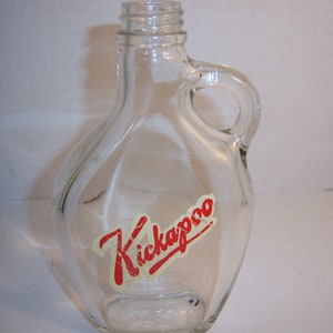 May include: Clear glass bottle with a handle and a red and white "Kickapoo" label. The bottle has a rounded shape and a spout for pouring. The handle is a loop shape. The bottle is empty.
