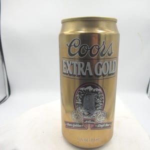 May include: A gold-colored Coors Extra Gold beer can. The can features the Coors logo in black script above the words "EXTRA GOLD". Below the text is a circular emblem with a mountain scene. The can is 12 fl oz (355 ml).