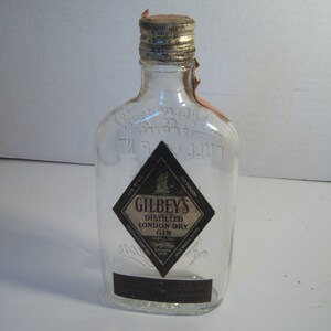 May include: A clear glass bottle of Gilbey's London Dry Gin. The bottle has a silver cap and a black and white label with the brand name and product information. The bottle is empty.