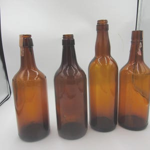 May include: Four empty amber glass bottles standing in a row. Each bottle features a narrow neck and a cylindrical body. Two of the bottles have small, light-colored paper remnants attached to the top of their necks, while the others are clear.