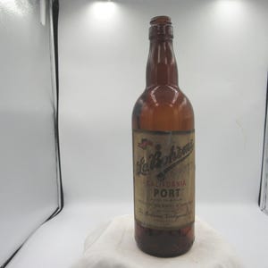 May include: A vintage brown glass bottle with a label that reads "La Paloma California Port." The bottle has a long neck and a rounded body. The label is aged and features text and decorative elements.