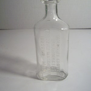 May include: A clear, antique glass bottle with a rectangular body and a rounded shoulder. The bottle has a narrow neck and a small, clear stopper. The text "WATER L. CONVER ST. PHARMACY BOSTON MASS" is embossed on the front.