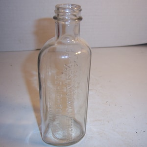 May include: A clear glass bottle with a textured surface. The bottle has a narrow neck and a wide base. The bottle is empty and has no label.