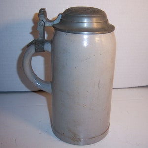 May include: A vintage stoneware beer stein with a hinged metal lid and handle. The cylindrical body is a light beige color, and the lid and handle hardware are silver. The stein has a classic, traditional design.