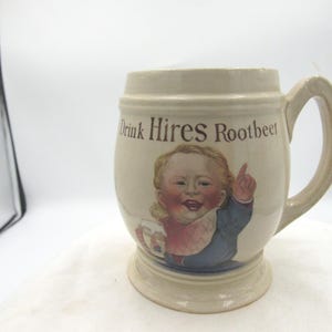 May include: Cream-colored ceramic mug with a handle, featuring the text "Drink Hires Rootbeer" and a smiling illustration of a child. The child is wearing a blue jacket and a bib. The mug has a wide base.