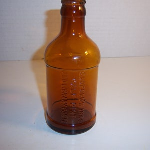 May include: Amber-colored glass bottle with a rounded body and a narrow neck. The bottle has embossed text that reads "SANFORD'S INKS HALF PINT AND LIBRARY PASTE". The bottle is empty and sits on a white surface.