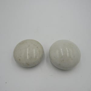 May include: Two smooth, oval-shaped, off-white stones are displayed against a white background. The stones have subtle variations in color and texture, with a slightly speckled appearance. They appear to be natural, possibly polished, stones.