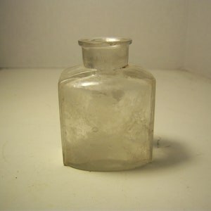May include: A clear, antique glass bottle with a rectangular body and a short, cylindrical neck. The bottle has a slightly textured surface and appears to be empty. The design is simple and functional, suggesting it may have been used for medicine or other liquids.