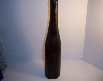 May include: A tall, dark brown glass bottle with a long, slender neck and a slightly wider base. The bottle appears to be empty and is likely designed for wine or other beverages. The bottle is standing upright.