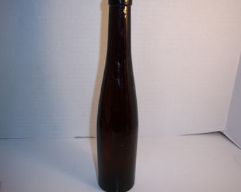 May include: A tall, brown glass bottle with a long neck and a rounded body. The bottle is empty and appears to be made of dark amber-colored glass. The bottle is approximately 12 inches tall.