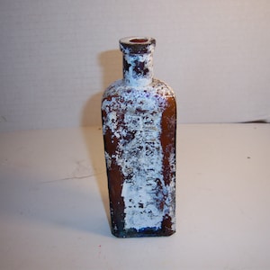 May include: A vintage glass bottle with a square shape and a frosted white finish. The bottle is brown with a blue tint and has a narrow neck.