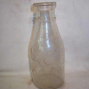 May include: A clear glass milk bottle with a textured surface. The bottle has a narrow neck and a wide base. The bottle is slightly dirty and has some scratches.