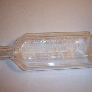 May include: A clear, rectangular glass bottle with a narrow neck and a wider body. The bottle has embossed text that reads "A.F. HOWARTH, CIR. COURT & WARWICK STS, UTICA, N.Y." The bottle is lying horizontally.