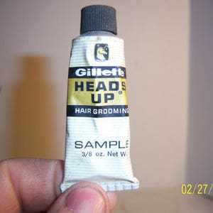 May include: A white tube of Gillette Heads Up Hair Grooming product with a black cap. The tube has a gold label with the text "Heads Up" and "Hair Grooming". The tube is labeled "Sample" and "3/8 oz. Net Wt."