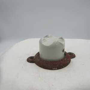 May include: A vintage electrical insulator with a white ceramic cylinder and a rusty red metal base. The base has two circular mounting holes. The insulator is likely from an older electrical system.