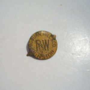May include: A vintage, circular pin with the text "RUGBURN, HILL & CO" and "R&W WINSTON" in a dark font on a yellow background. The pin has a small, pointed tab on the left side and a broken tab on the right.