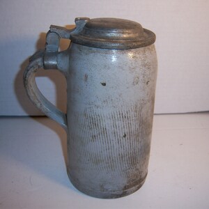 May include: A white ceramic beer stein with a pewter lid and a handle. The stein has a ribbed design and is slightly weathered.