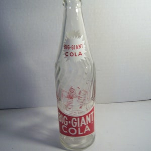 May include: Clear glass Big Giant Cola bottle with red and white labels. The labels feature the brand name and a graphic of a soda fountain. The bottle has a unique, ridged design.